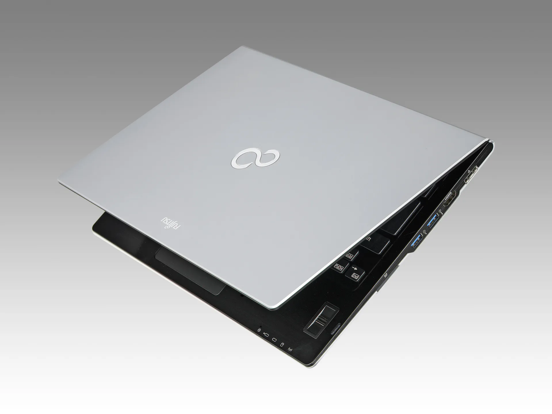 Fujitsu LifeBook U772_0306_03 - lab-kuzniewski.pl - Testy laptopów, recenzje i porównania Angled view of a silver laptop opened to reveal part of the keyboard and side ports against a gray gradient background.