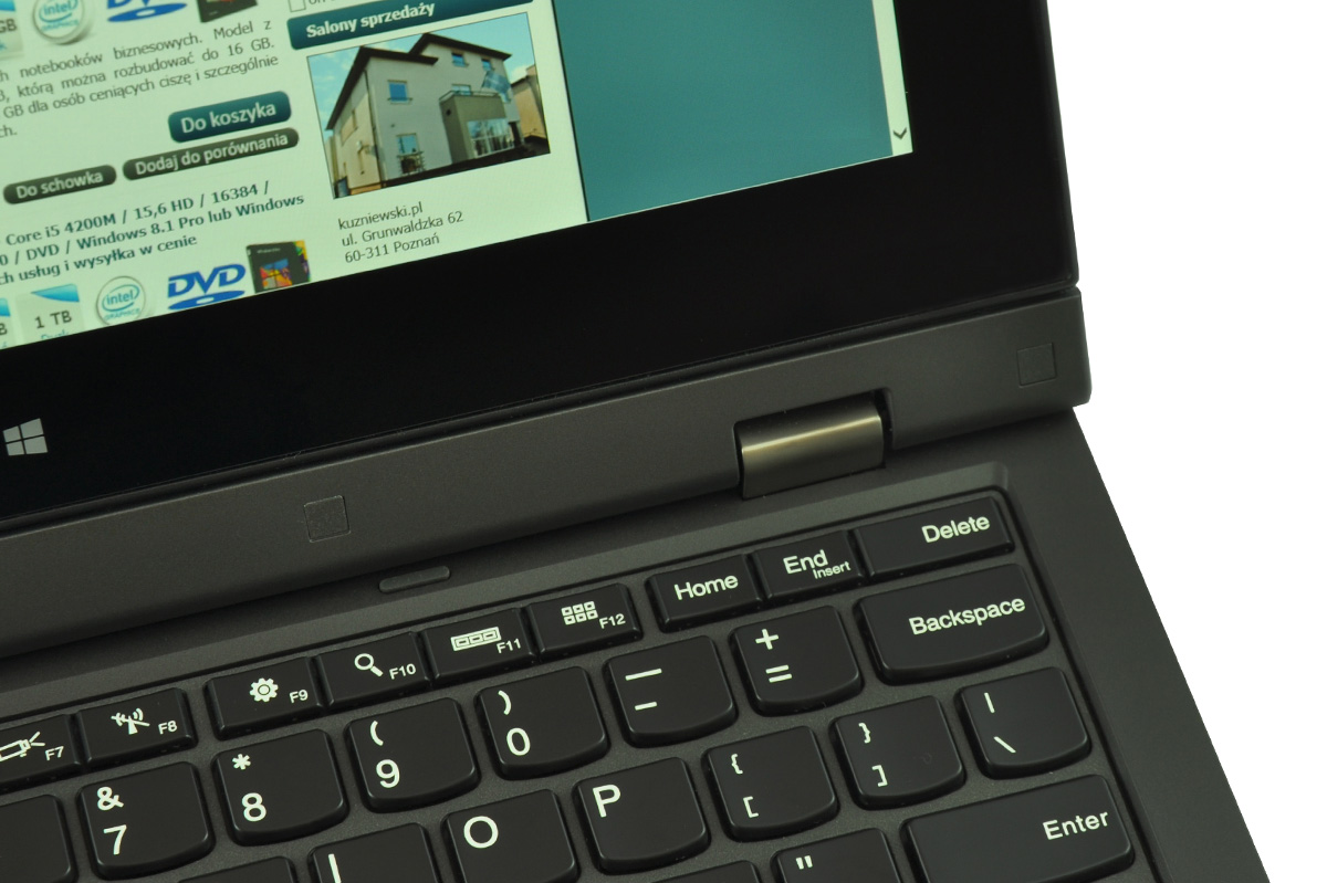 Close-up of a black laptop keyboard and hinge with a screen showing a Polish web page featuring a house image and buttons like Do koszyka and Do schowka.