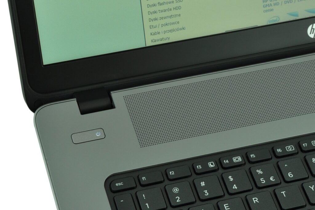Close-up of a gray HP laptop's palmrest: power button left of the keyboard and a perforated speaker grille, with part of the screen visible above.