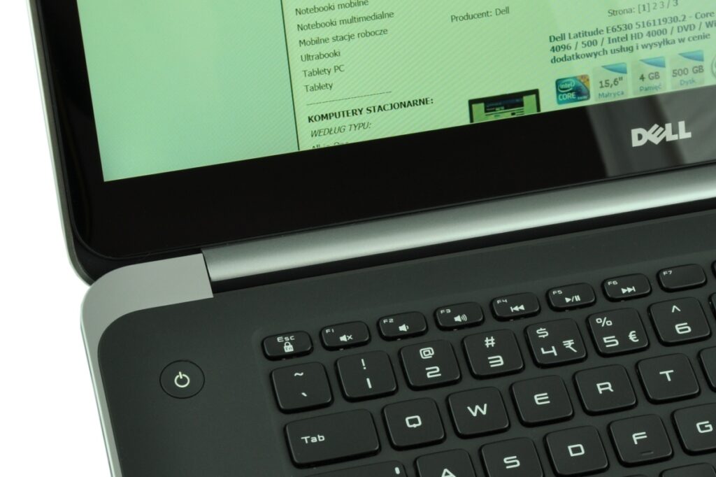 Close-up of a Dell laptop showing the left side of the keyboard and a power button, with a green monitor displaying text on screen in the background
