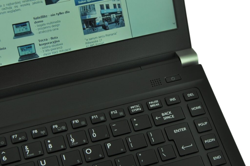 Close-up of a black laptop showing the keyboard and a portion of the display; the screen shows a Polish webpage with text and images, and a power button is visible near the hinge.