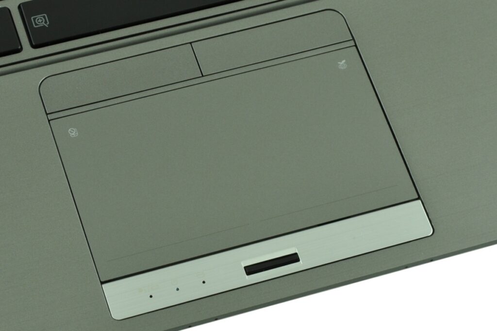 Close-up of a laptop touchpad area with a large, recessed trackpad, a slim slot beneath it, and small indicator holes on the metal base plate