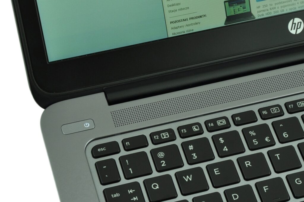 Close-up of a gray HP laptop keyboard, showing the power button on the left, a perforated speaker grille above the keys, and part of the screen with an HP logo on the bezel.