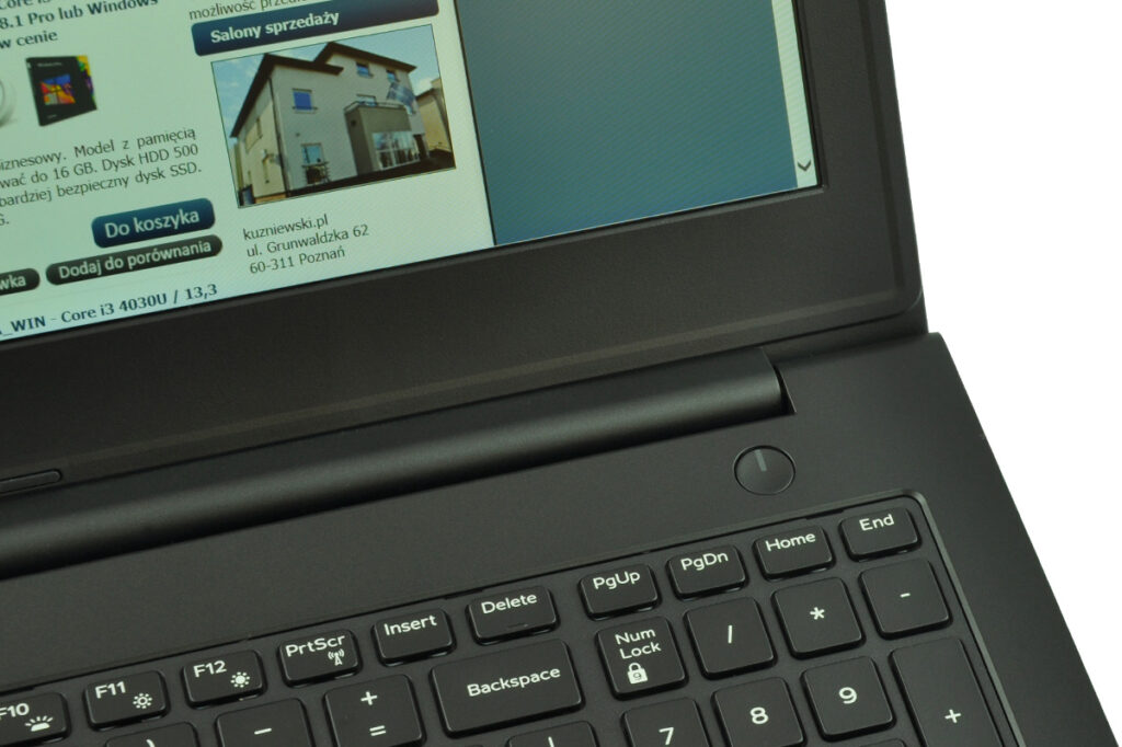 Close-up of a dark laptop with a keyboard; the screen shows a Polish website with a house photo and blue/green layout.