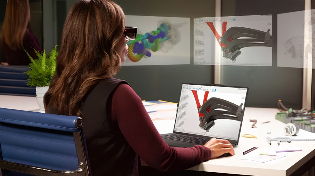 Engineer wearing a headset examines a CAD model of a robotic gripper on her laptop at a cluttered white desk.