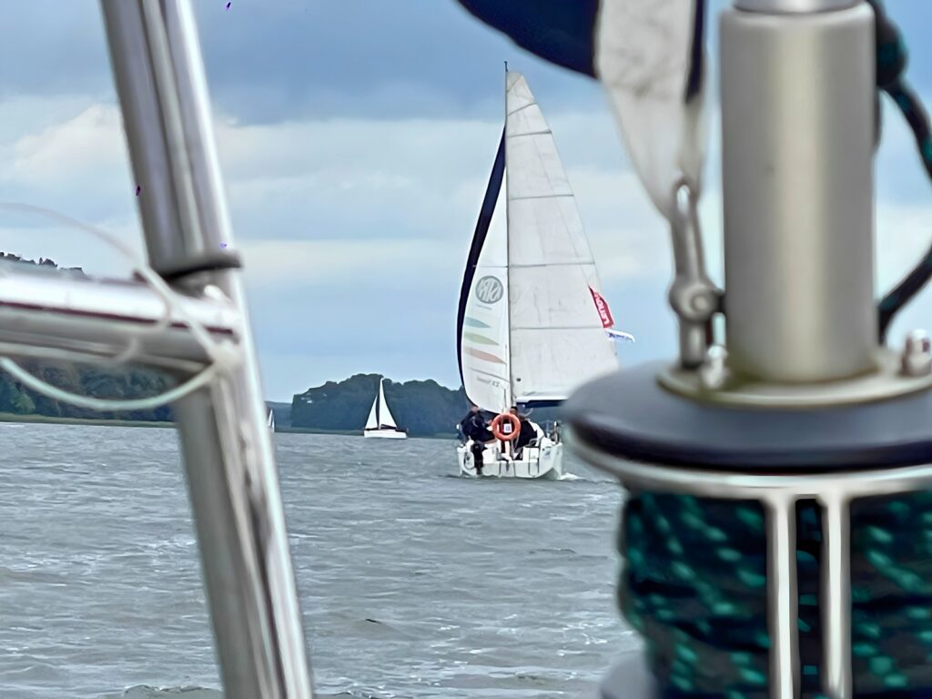 Sailboat with a tall white sail and an orange life ring on its stern, sailing on choppy water framed by boat rigging in the foreground.