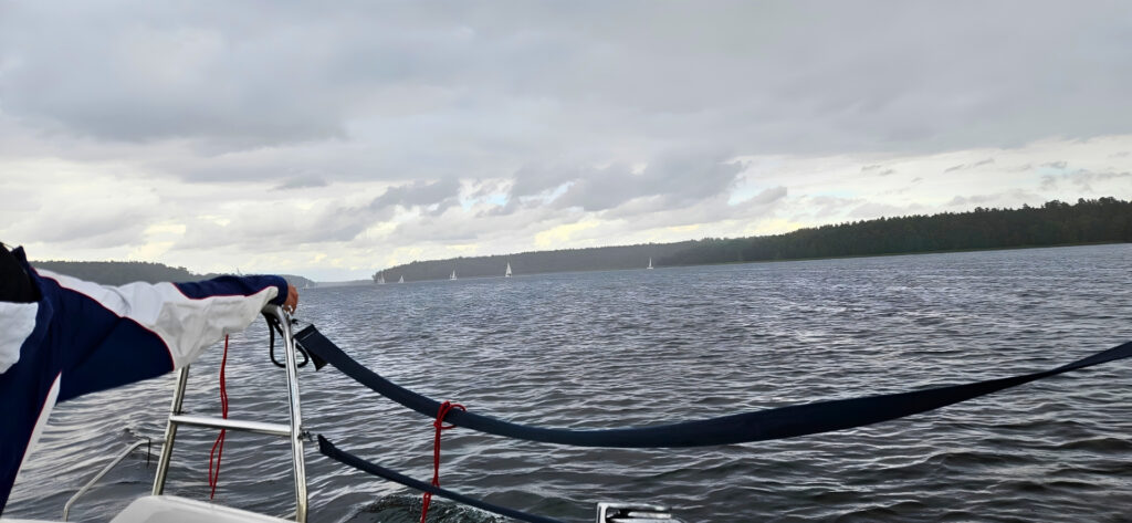 View from a sailboat deck: a person’s arm in a blue–white jacket grips the railing while a cloudy lake and distant sailboats unfold ahead.
