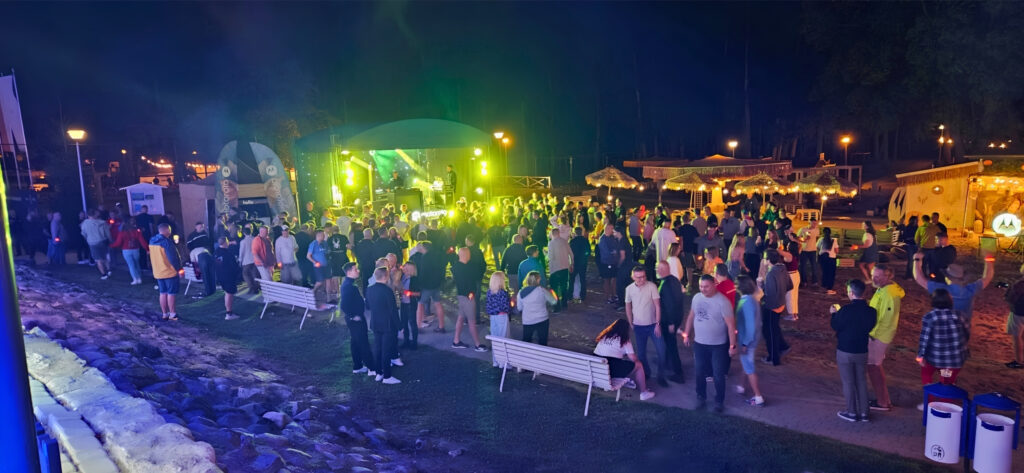 Crowd at a nighttime outdoor concert with a green-lit stage and tents in the background.