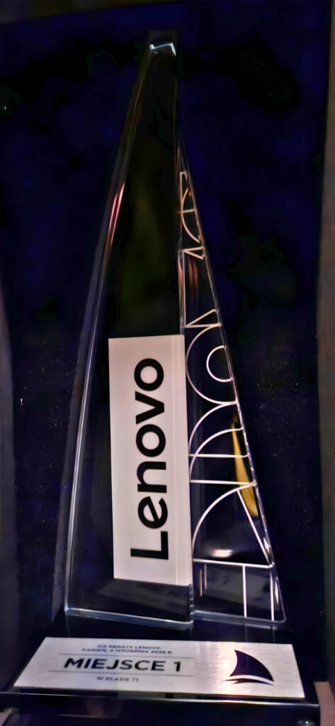 Tall glass award with a sail-like triangular shape inside a dark display case; a white placard at the base reads 'MIEJSCE 1' (first place) with a small sailboat logo nearby.