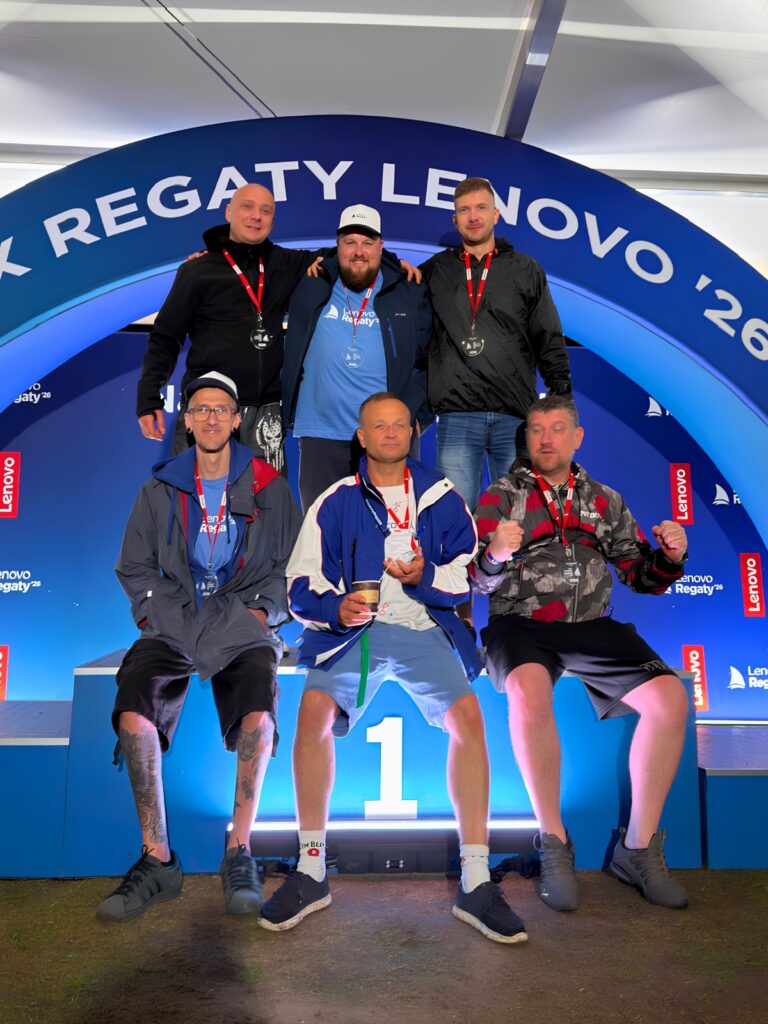 Six men with medals posing on a first-place podium under a blue arch at a Lenovo Regaty event.