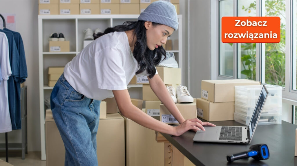 Woman in a white t-shirt and beanie typing on a laptop at a storage workspace with shoe boxes on shelves behind her and on the desk