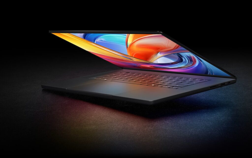 Slim gray laptop opened at a shallow angle on a dark surface, glowing multicolor screen lighting the scene.