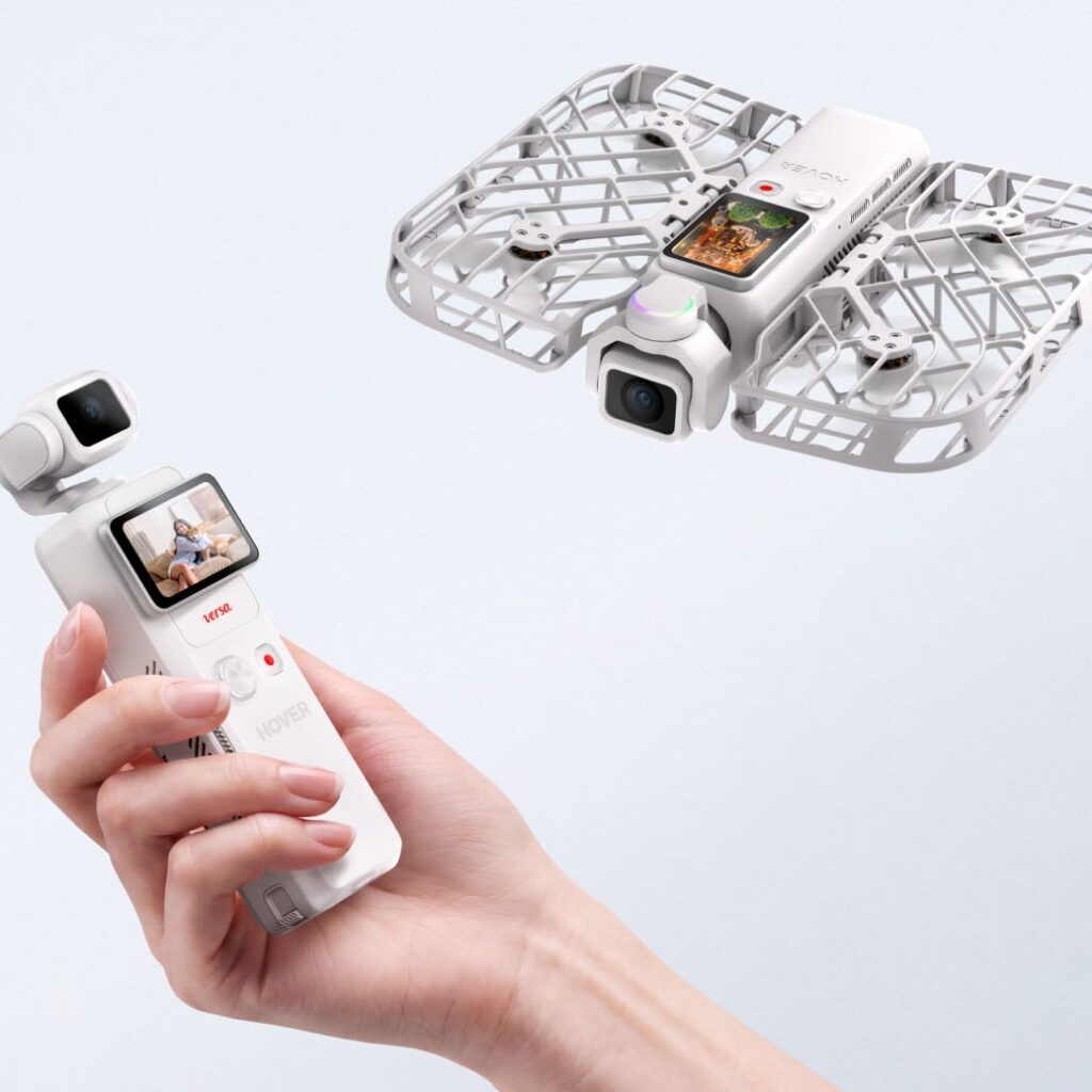 Hand holding a white drone remote/controller with a small screen, while a white quadcopter with a camera hovers nearby above.