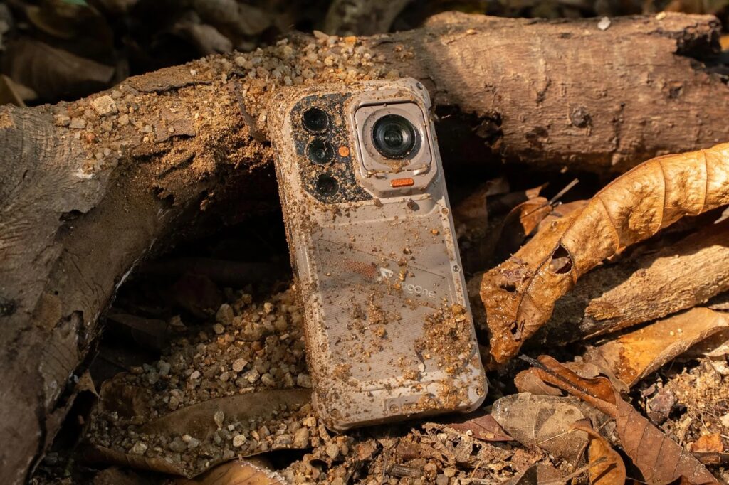 Rugged smartphone covered in dirt and mud lying among logs and dry leaves on the forest floor.